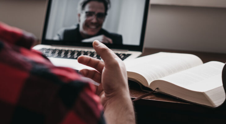 Person in red plaid shirt gestures during a video call on a laptop, with a smiling man on screen. An open book lies on the desk near the hand.