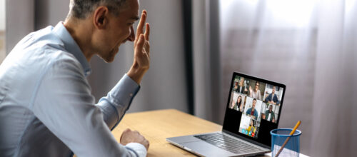 A man sitting at a desk raises his hand to ask a question during a video conference call