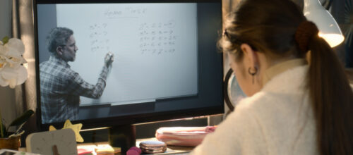 A student watches a professor on a computer screen and takes notes