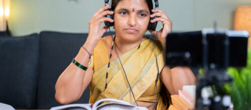 A woman sitting in front of a camera and some books puts a pair of headphones on