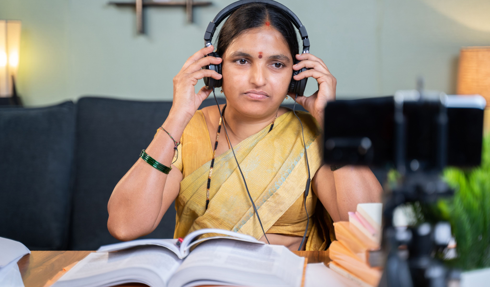 A woman sitting in front of a camera and some books puts a pair of headphones on