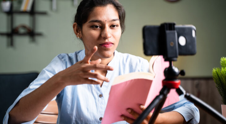 A woman reading from a book and recording herself with her phone gestures toward the camera
