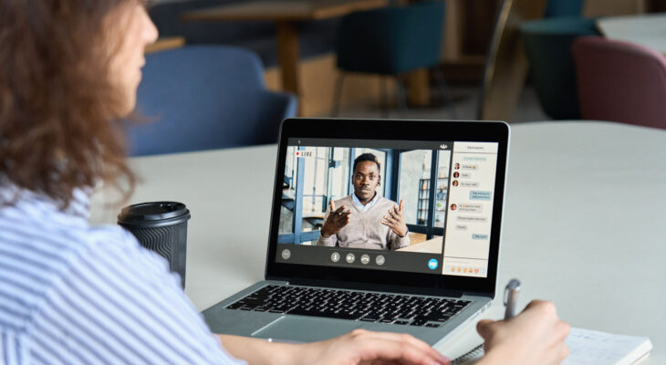 A woman takes notes as she watches a man on a video call give a presentation