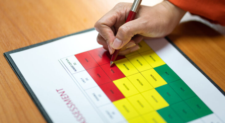 A hand holding a red pen fills out a colorful risk assessment chart, segmented in red, yellow, and green. The focus is on high-risk red areas.