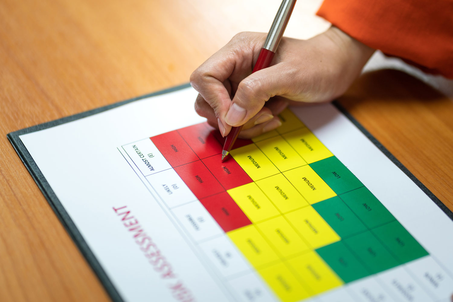 A hand holding a red pen fills out a colorful risk assessment chart, segmented in red, yellow, and green. The focus is on high-risk red areas.