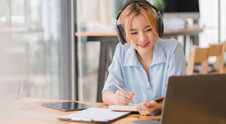 A woman smiles while looking at her laptop and taking notes