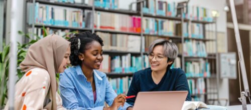 A group of students in a library sit around a laptop and talk