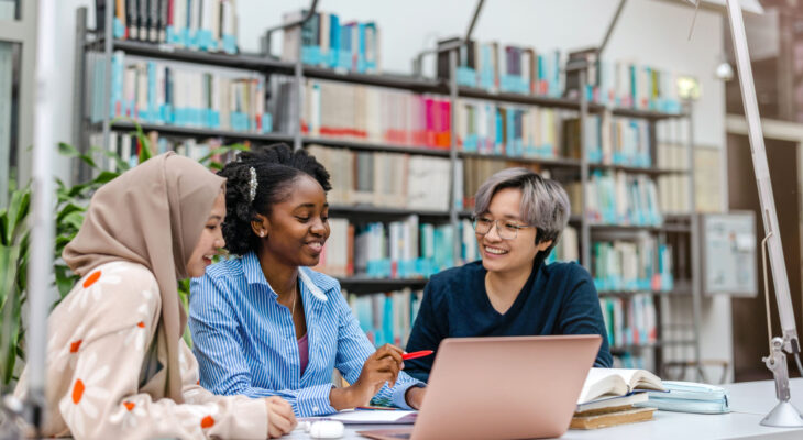 A group of students in a library sit around a laptop and talk