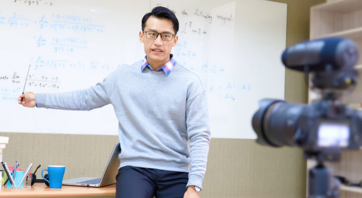 A professor standing in front of a white board speaks into a camera