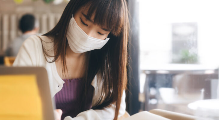 Young adult asian female with protective face mask is working on a laptop at a cafe.