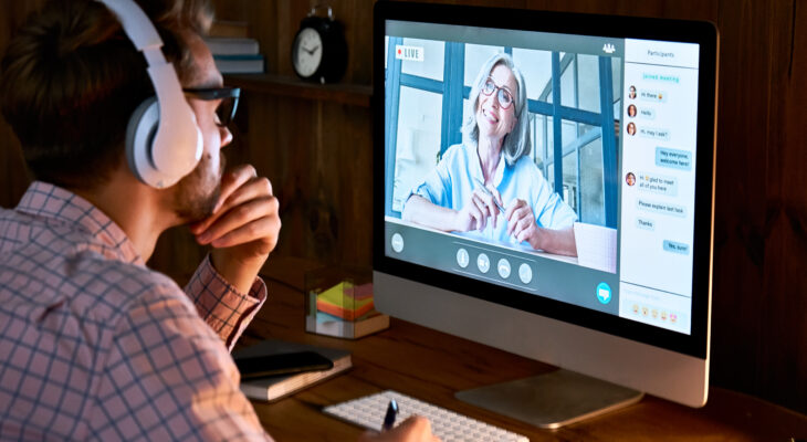 Male student wearing headphones taking online course seminar, watching webinar featuring a woman.