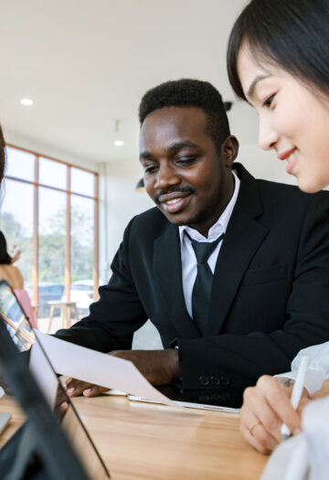 Three diverse professionals collaborate at a table in a bright office space. A man in a black suit is holding a document, while two women, one in a yellow blazer and the other in a white blouse, smile and engage with the content on a laptop and tablet.