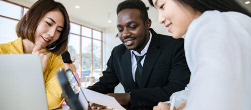 Three diverse professionals collaborate at a table in a bright office space. A man in a black suit is holding a document, while two women, one in a yellow blazer and the other in a white blouse, smile and engage with the content on a laptop and tablet.