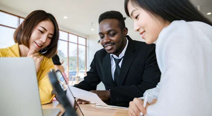 Three diverse professionals collaborate at a table in a bright office space. A man in a black suit is holding a document, while two women, one in a yellow blazer and the other in a white blouse, smile and engage with the content on a laptop and tablet.