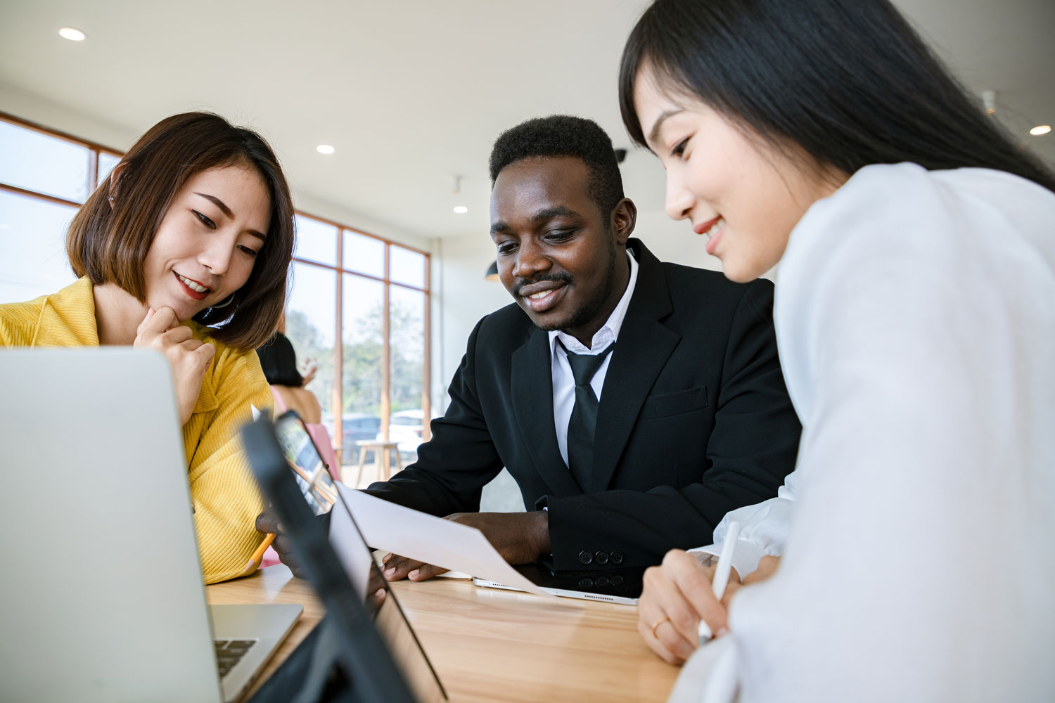 Three diverse professionals collaborate at a table in a bright office space. A man in a black suit is holding a document, while two women, one in a yellow blazer and the other in a white blouse, smile and engage with the content on a laptop and tablet.