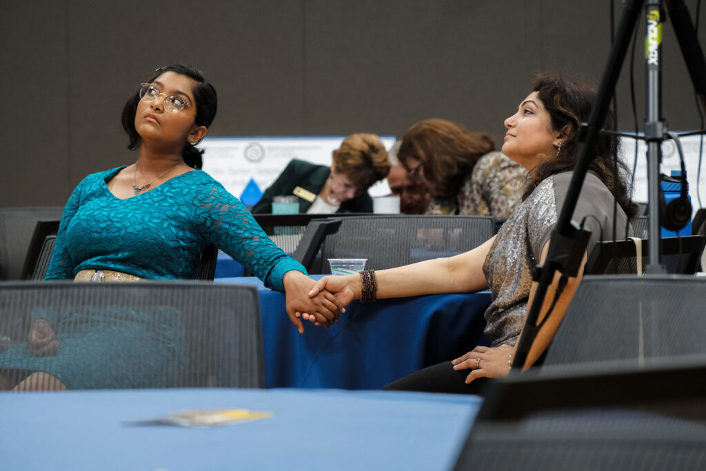 A young girl in a teal dress and an older woman in a silver top hold hands across a table, both gazing upward with intense expressions. Other people are seated in the background, focused downward.