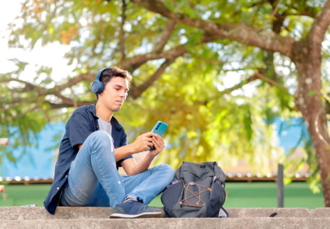 A person is sitting on outdoor steps, wearing headphones and holding a smartphone. They have on a dark blue shirt, light blue jeans, and black slip-on shoes. Next to them is a black backpack with visible zipper compartments and orange string loops. The background shows green foliage with sunlight filtering through the leaves, and there is a blue section of a building partially visible behind the trees.