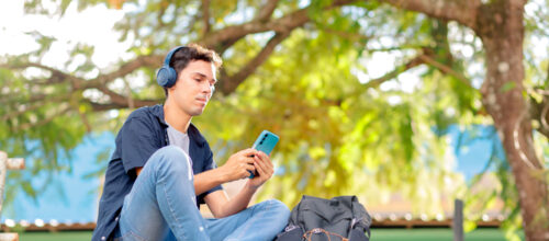 A person is sitting on outdoor steps, wearing headphones and holding a smartphone. They have on a dark blue shirt, light blue jeans, and black slip-on shoes. Next to them is a black backpack with visible zipper compartments and orange string loops. The background shows green foliage with sunlight filtering through the leaves, and there is a blue section of a building partially visible behind the trees.
