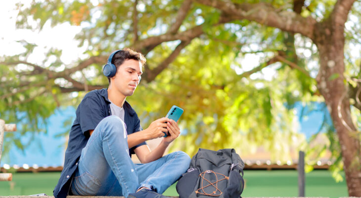 A person is sitting on outdoor steps, wearing headphones and holding a smartphone. They have on a dark blue shirt, light blue jeans, and black slip-on shoes. Next to them is a black backpack with visible zipper compartments and orange string loops. The background shows green foliage with sunlight filtering through the leaves, and there is a blue section of a building partially visible behind the trees.