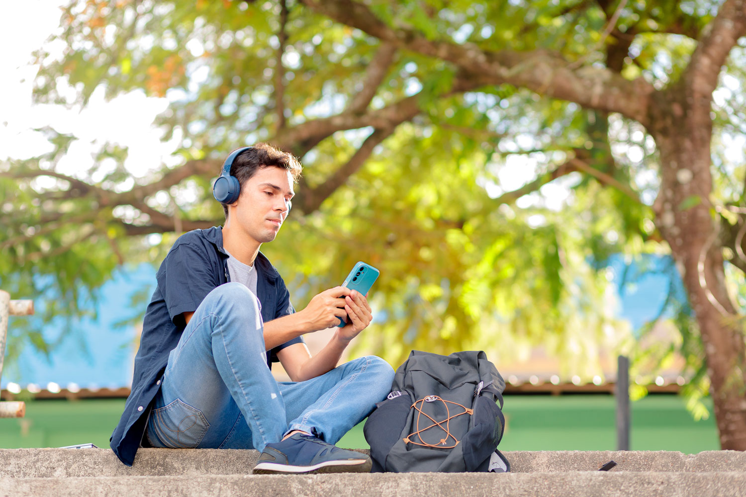 A person is sitting on outdoor steps, wearing headphones and holding a smartphone. They have on a dark blue shirt, light blue jeans, and black slip-on shoes. Next to them is a black backpack with visible zipper compartments and orange string loops. The background shows green foliage with sunlight filtering through the leaves, and there is a blue section of a building partially visible behind the trees.