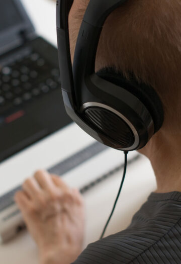 A person wearing large over-ear headphones is focused on a laptop, with one hand on a Braille display in front of the keyboard.
