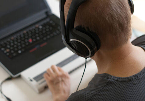 A person wearing large over-ear headphones is focused on a laptop, with one hand on a Braille display in front of the keyboard.