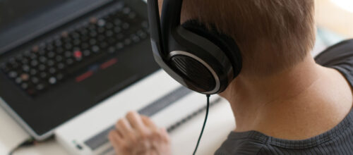 A person wearing large over-ear headphones is focused on a laptop, with one hand on a Braille display in front of the keyboard.