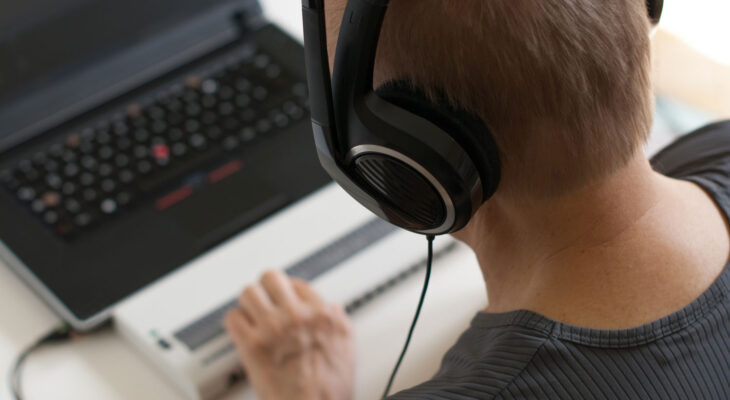 A person wearing large over-ear headphones is focused on a laptop, with one hand on a Braille display in front of the keyboard.