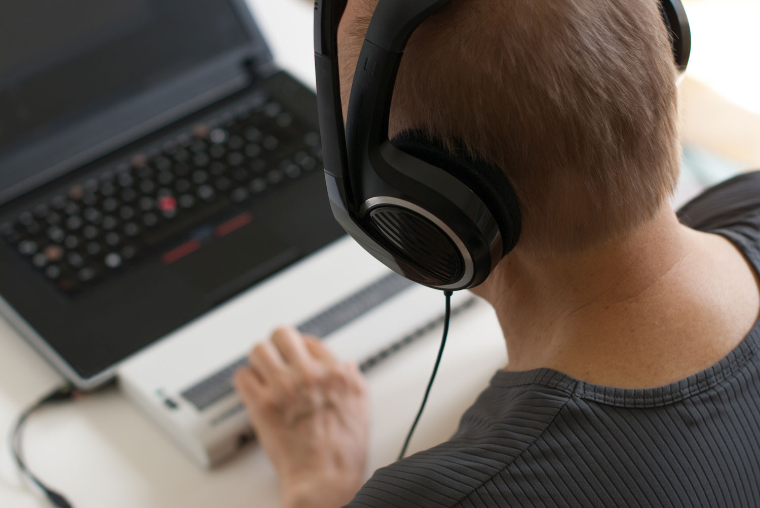 A person wearing large over-ear headphones is focused on a laptop, with one hand on a Braille display in front of the keyboard.