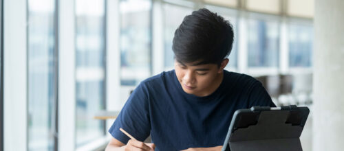 A student sits at a table near tall windows, writing in a notebook with a tablet propped open beside them.