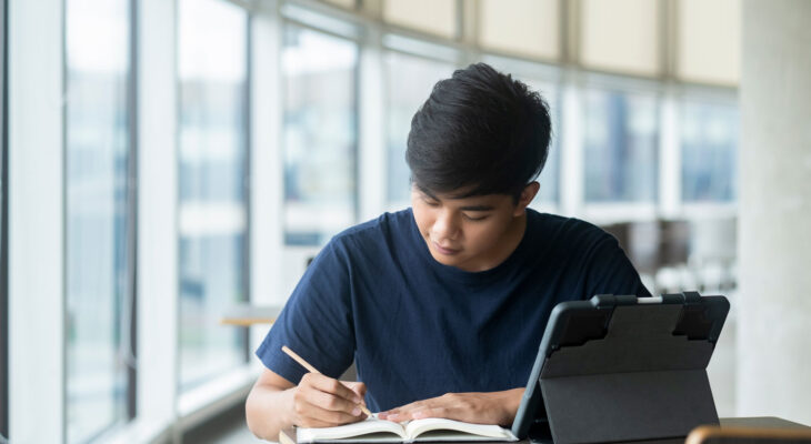 A student sits at a table near tall windows, writing in a notebook with a tablet propped open beside them.
