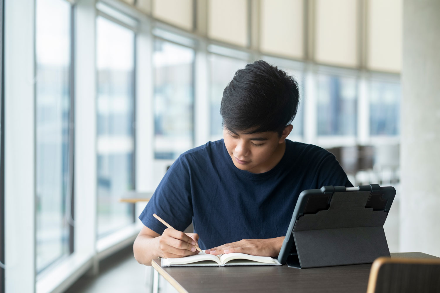 A student sits at a table near tall windows, writing in a notebook with a tablet propped open beside them.