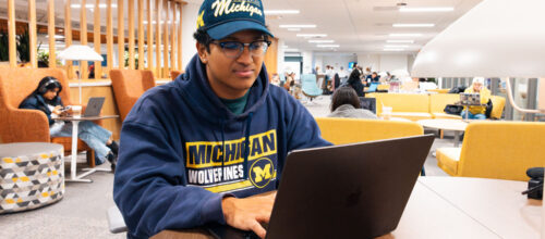 Student in Michigan Wolverines gear works on a laptop in a busy, modern university study lounge.