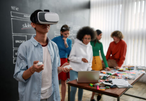 A student stands at the front wearing a virtual reality headset and holding controllers. Behind him, five peers gather around a table with a laptop and colorful supplies, suggesting a collaborative project or creative workshop.