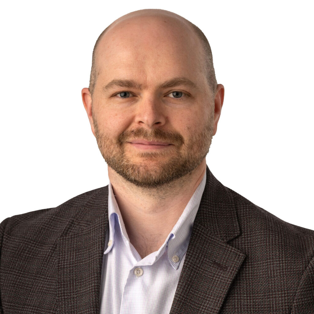 A head-and-shoulders portrait photograph of a bad man with brown beard wearing a dark sportcoat and light collared shirt.