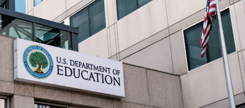 Exterior sign of the U.S. Department of Education building with its seal and a U.S. flag on a pole in the foreground.