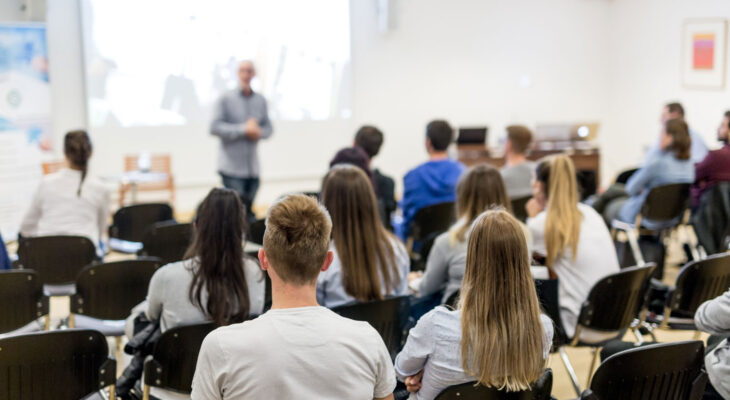 Students are seated in rows facing a speaker at the front of a classroom, with a presentation projected behind him.