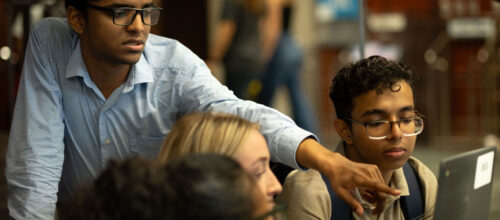 A group of students gathers around a laptop; one student in a light blue shirt points at the screen while another, wearing glasses and a backpack, looks on attentively.