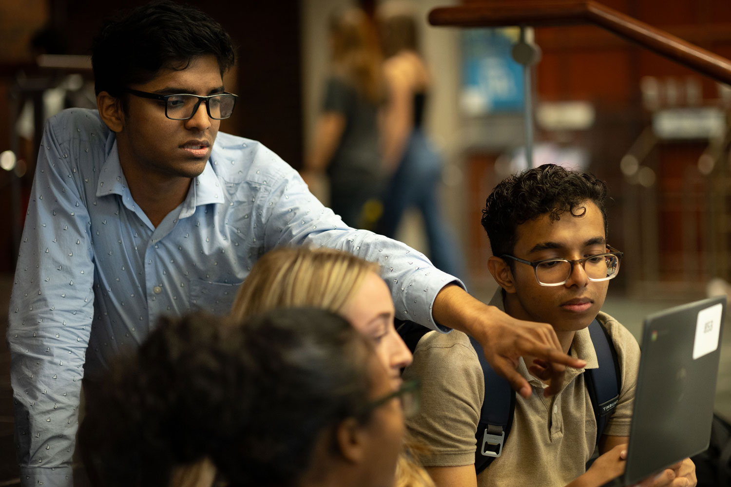 A group of students gathers around a laptop; one student in a light blue shirt points at the screen while another, wearing glasses and a backpack, looks on attentively.