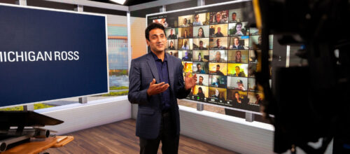 Professor lectures from a studio with Michigan Ross signage and a live Zoom wall of students in the background.