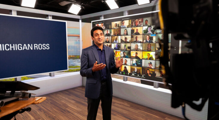 Professor lectures from a studio with Michigan Ross signage and a live Zoom wall of students in the background.