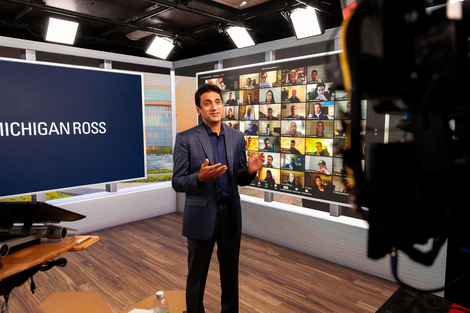 Professor lectures from a studio with Michigan Ross signage and a live Zoom wall of students in the background.