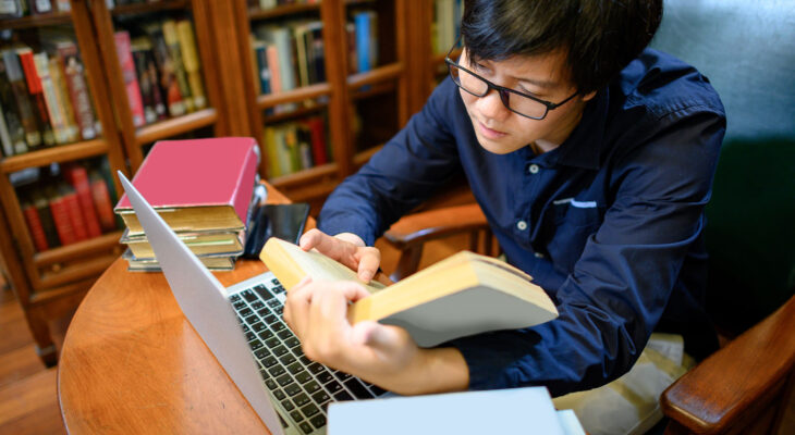 A young adult wearing glasses studies in a library, reading a book while using a laptop. Several books are stacked on the table.