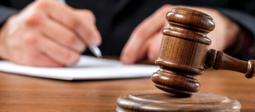 A wooden judge’s gavel rests on a desk while a person in a suit writes on paper in the blurred background.