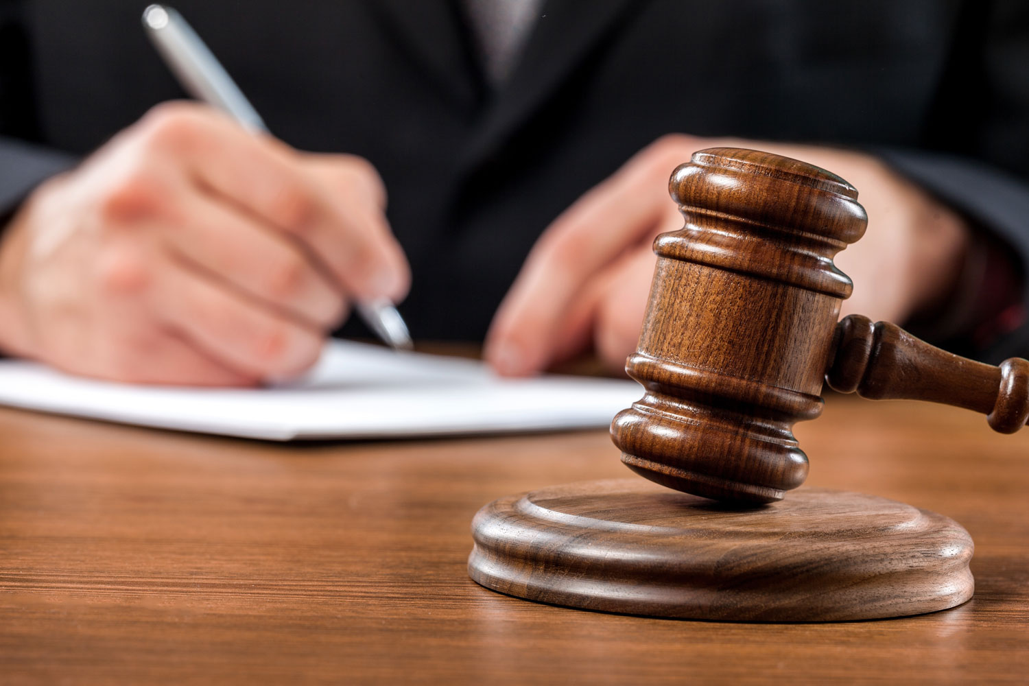 A wooden judge’s gavel rests on a desk while a person in a suit writes on paper in the blurred background.