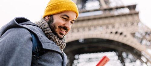 Smiling man in a yellow beanie looks at his phone with the Eiffel Tower in the background on a cloudy day.