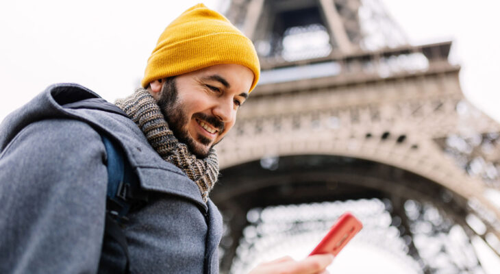 Smiling man in a yellow beanie looks at his phone with the Eiffel Tower in the background on a cloudy day.
