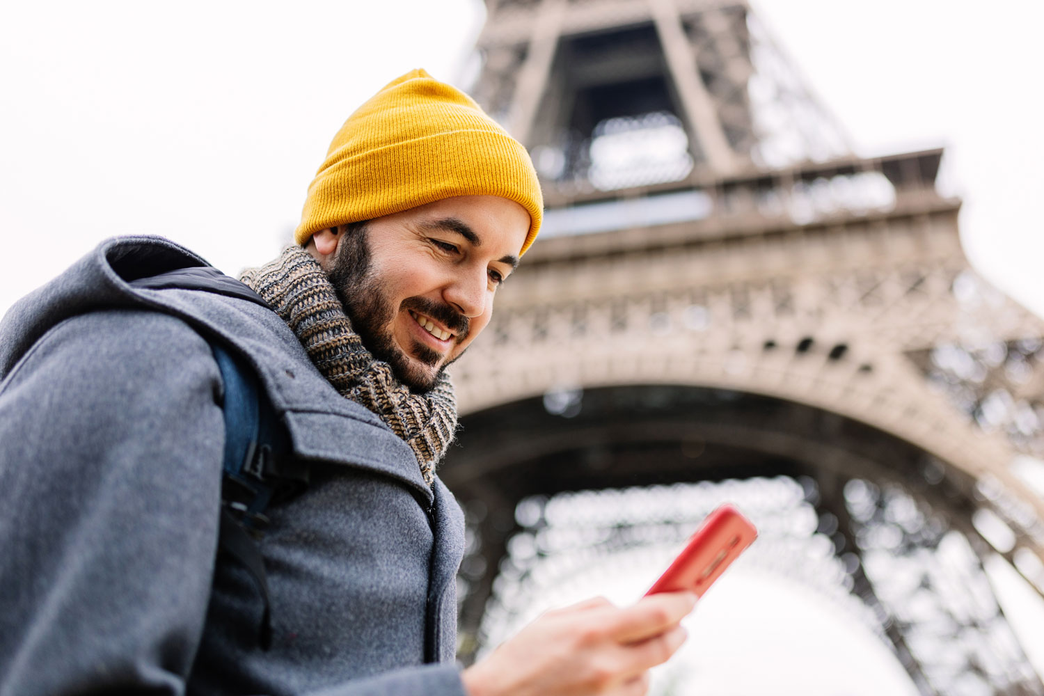 Smiling man in a yellow beanie looks at his phone with the Eiffel Tower in the background on a cloudy day.