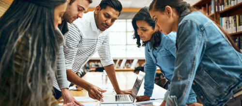 Five people lean over a table covered in documents, collaborating on a project in a well-lit library or office space.