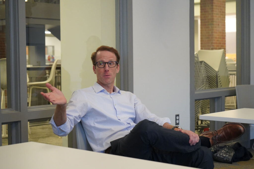 Man in glasses gestures while seated in discussion, with stacked chairs and windows in the background.